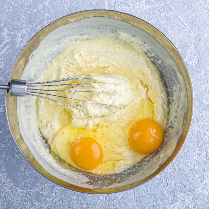 Glass mixing bowl with two egg yolks, whisk, and cake batter, top view with bowl on blue table
