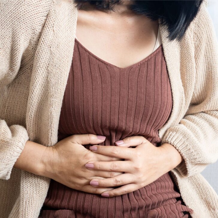 woman with a brown top and tan cardigan holding her stomach - signs of estrogen dominance