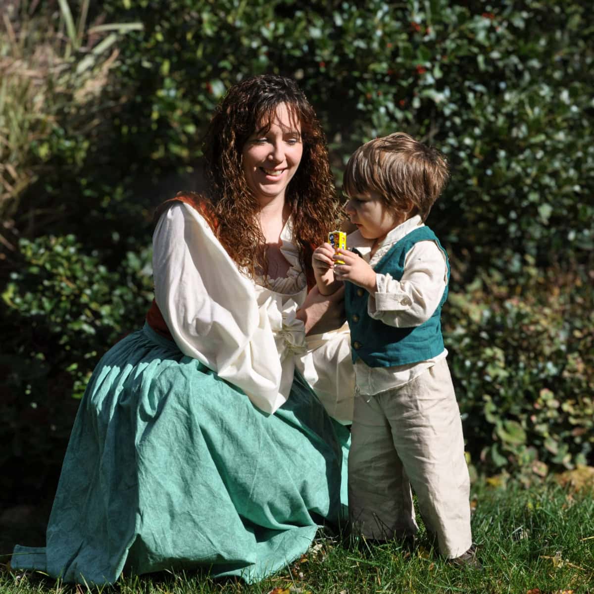 Bonnie and her young son Henry sitting together in a lush garden. Bonnie is wearing a flowing green skirt and rustic blouse, while Henry is dressed in a charming vest and linen outfit, holding a small toy. The sunlight highlights their warm smiles and the natural beauty around them, evoking a sense of connection, simplicity, and joy.