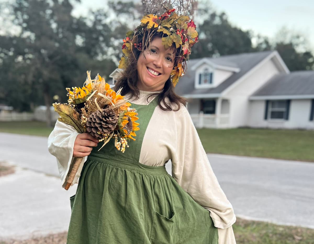 Bonnie dressed as a tree nymph for Halloween, wearing a flowing green pinafore and a crown of autumn leaves, holding a bouquet of sunflowers, pinecones, and natural elements. Set against a backdrop of a cozy neighborhood, this image reflects her connection to nature, creativity, and the whimsical charm that embodies her homesteading and homemaking journey.