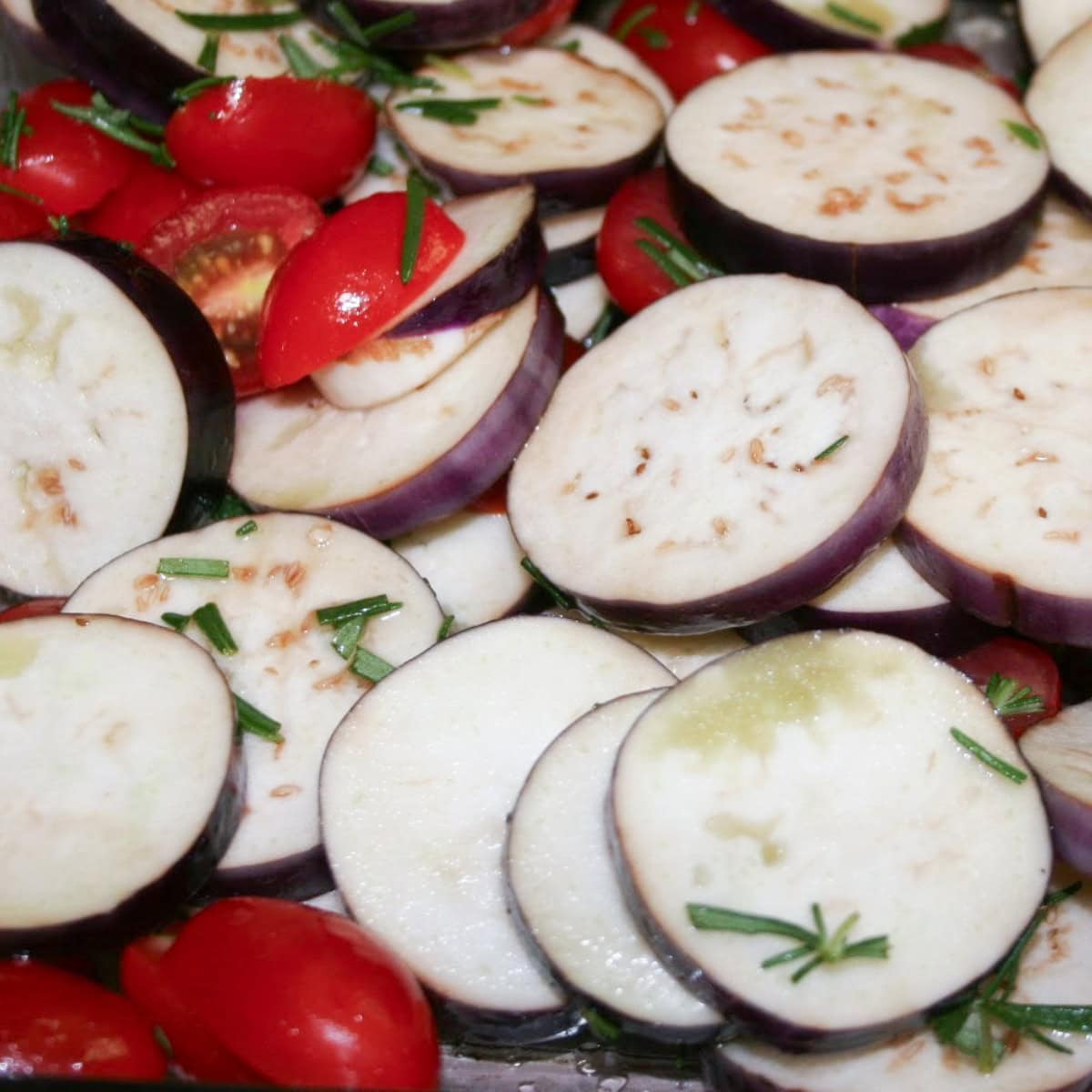 Freshly sliced eggplant and halved cherry tomatoes from the garden, sprinkled with chopped rosemary, ready to be roasted in the oven. A vibrant representation of homegrown produce being transformed into a delicious and wholesome dish, perfect for the Harvest to Table journey.