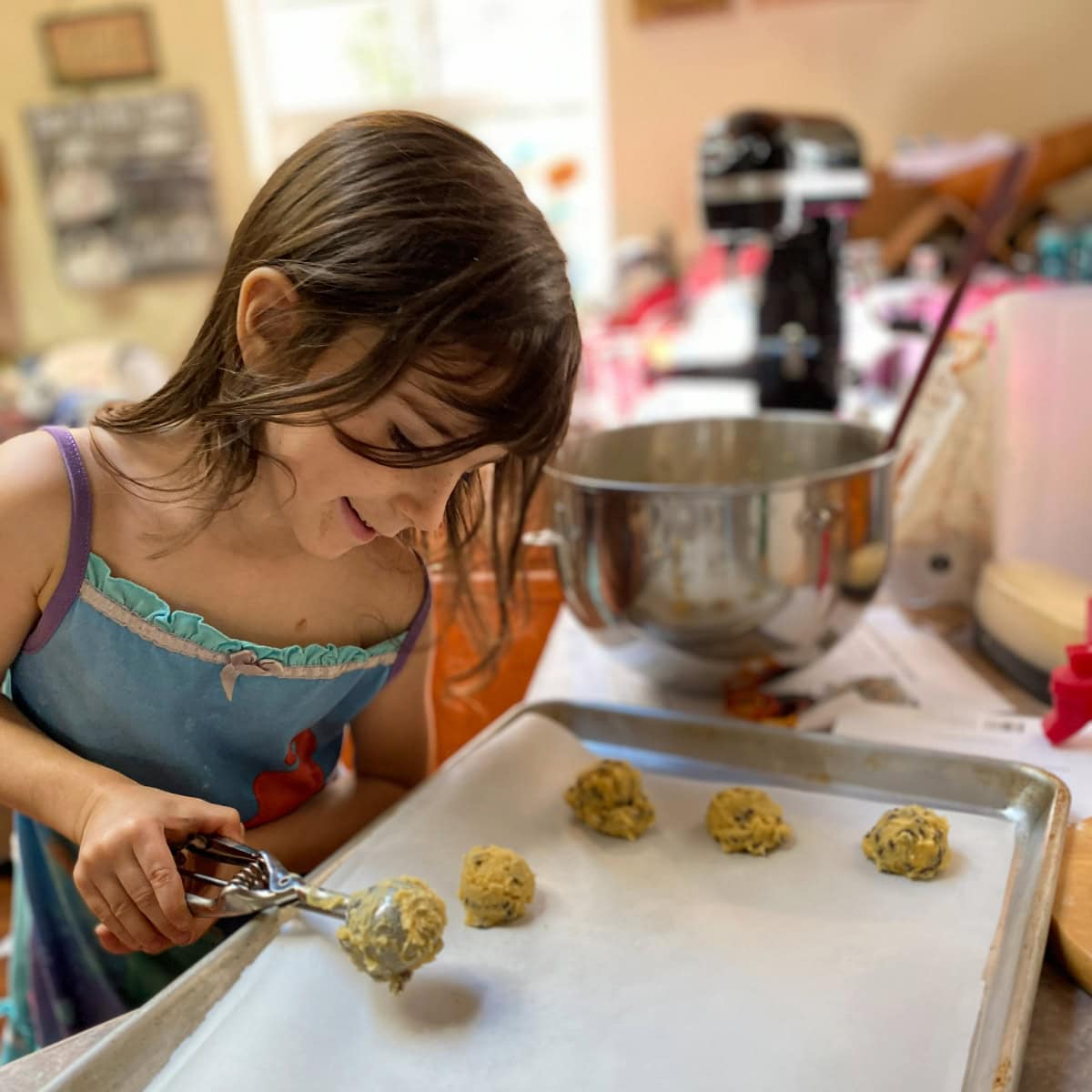 A joyful moment in the kitchen as Elise, a young girl, carefully scoops cookie dough onto a baking sheet. Surrounded by a mixing bowl and baking supplies, she smiles with focus and excitement, capturing the warmth and creativity of family baking time.