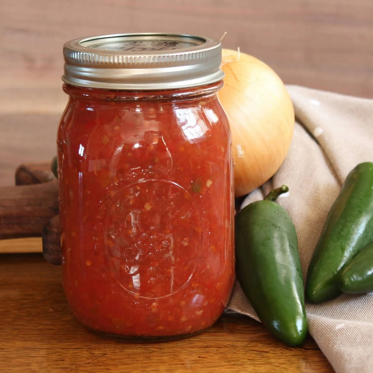 A jar of homemade canned salsa sits on a wooden surface, surrounded by fresh ingredients including jalapeños and an onion. The vibrant red salsa showcases the rich flavors of a garden-fresh recipe, perfect for preserving and enjoying year-round.